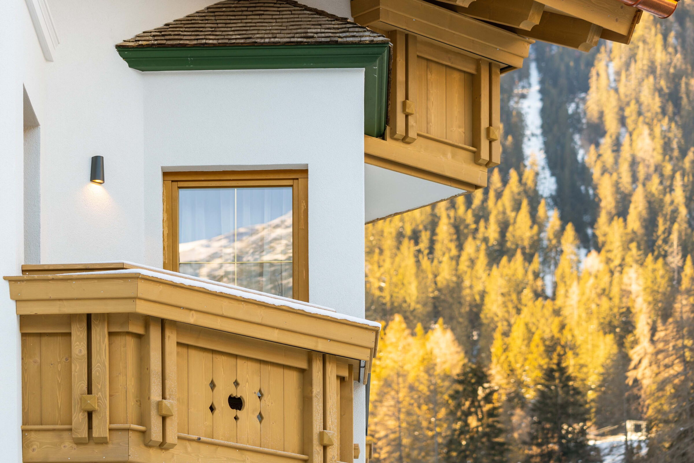 Aussenansicht der Dorfstube mit Holzverzierungen und Balkon, im Hintergrund Berge und Wald.