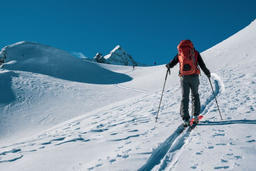 Skitourengeher mit rotem Rucksack steigt mit Tourenski und Stöcken in einer Spur über einen verschneiten Hang auf, im Hintergrund Gipfel unter blauem Himmel
